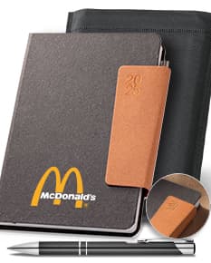 Agenda A5 com fecho magnético em couro marrom, vazia e aberta em uma mesa de escritório. Ao lado, há um notebook, carteira, estojo, papéis e dinheiro. A imagem revela um ambiente de negócios com objetos vintage e tecnológicos.