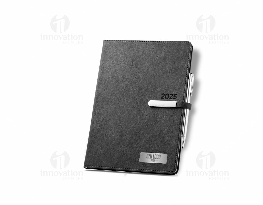 Agenda B5 com fecho magnético brilhante, em marrom de couro, aberta sobre uma mesa contendo um notebook, caderno de anotações, páginas em branco e uma carteira. Ao fundo, um teclado de computador e um pen drive.