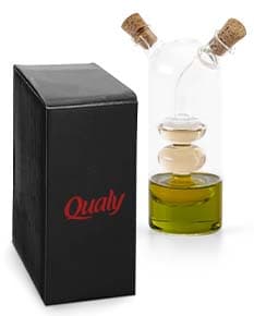 Galheteiro de vidro com três recipientes, um para azeite, outro para vinagre e um terceiro para molho de pimenta. Cada recipiente possui uma tampa de metal e um bico dosador. O galheteiro está sobre uma mesa de madeira escura.