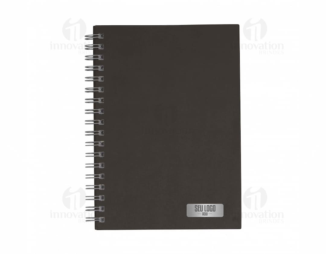 Caderno em branco aberto sobre uma mesa de escritório, com uma caneta ao lado. O caderno possui linhas para escrever e um design simples e moderno. A imagem também mostra um notebook e outros materiais de escrita ao fundo.