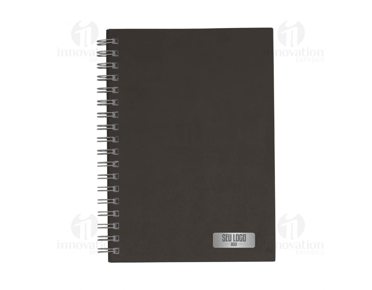 Caderno em branco aberto sobre uma mesa de escritório, com uma caneta ao lado. O caderno possui linhas para escrever e um design simples e moderno. A imagem também mostra um notebook e outros materiais de escrita ao fundo.