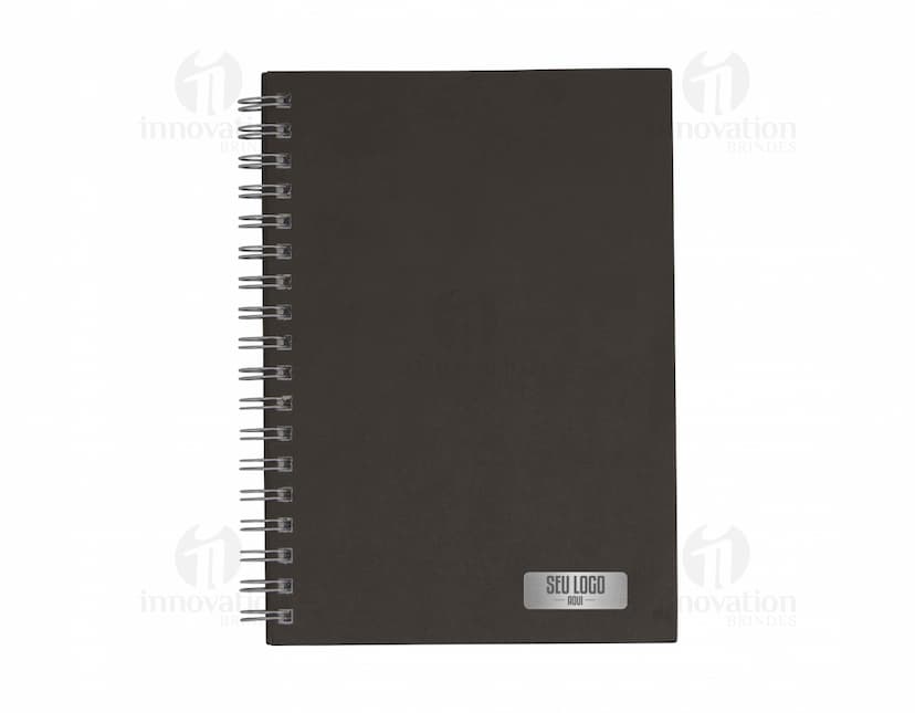 Caderno em branco aberto sobre uma mesa de escritório, com uma caneta ao lado. O caderno possui linhas para escrever e um design simples e moderno. A imagem também mostra um notebook e outros materiais de escrita ao fundo.