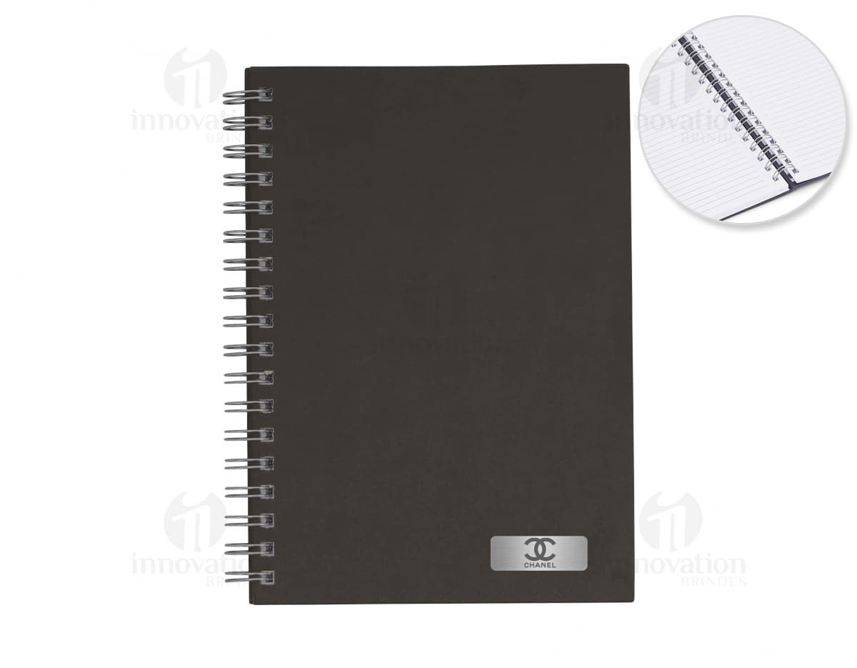 Caderno em branco aberto sobre uma mesa de escritório, com uma caneta ao lado. O caderno possui linhas para escrever e um design simples e moderno. A imagem também mostra um notebook e outros materiais de escrita ao fundo.