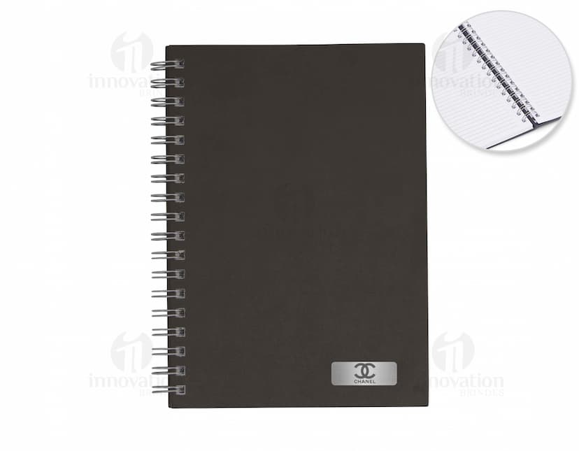 Caderno em branco aberto sobre uma mesa de escritório, com uma caneta ao lado. O caderno possui linhas para escrever e um design simples e moderno. A imagem também mostra um notebook e outros materiais de escrita ao fundo.