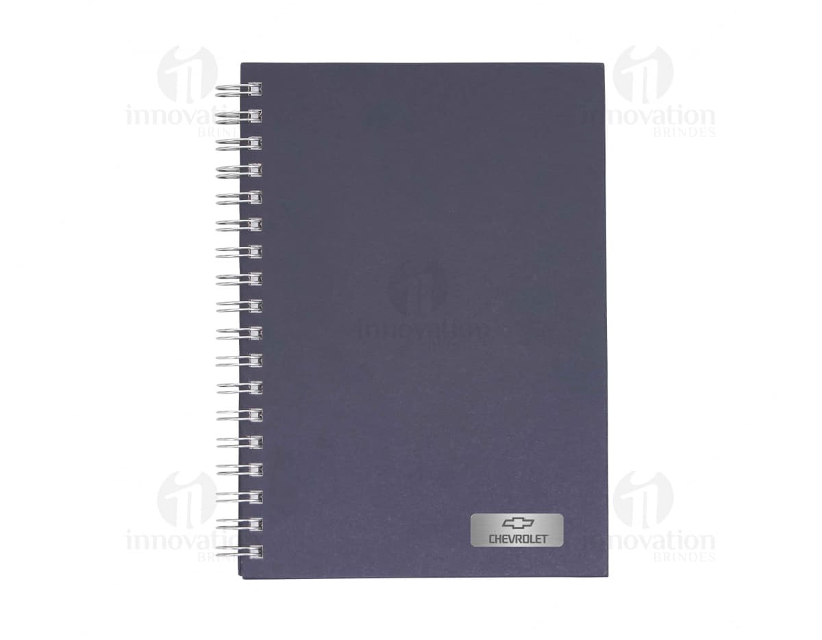 Caderno em branco aberto sobre uma mesa de escritório, com uma caneta ao lado. O caderno possui linhas para escrever e um design simples e moderno. A imagem também mostra um notebook e outros materiais de escrita ao fundo.