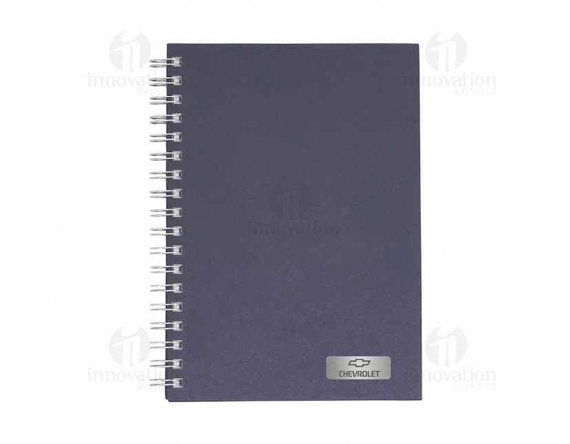Caderno em branco aberto sobre uma mesa de escritório, com uma caneta ao lado. O caderno possui linhas para escrever e um design simples e moderno. A imagem também mostra um notebook e outros materiais de escrita ao fundo.