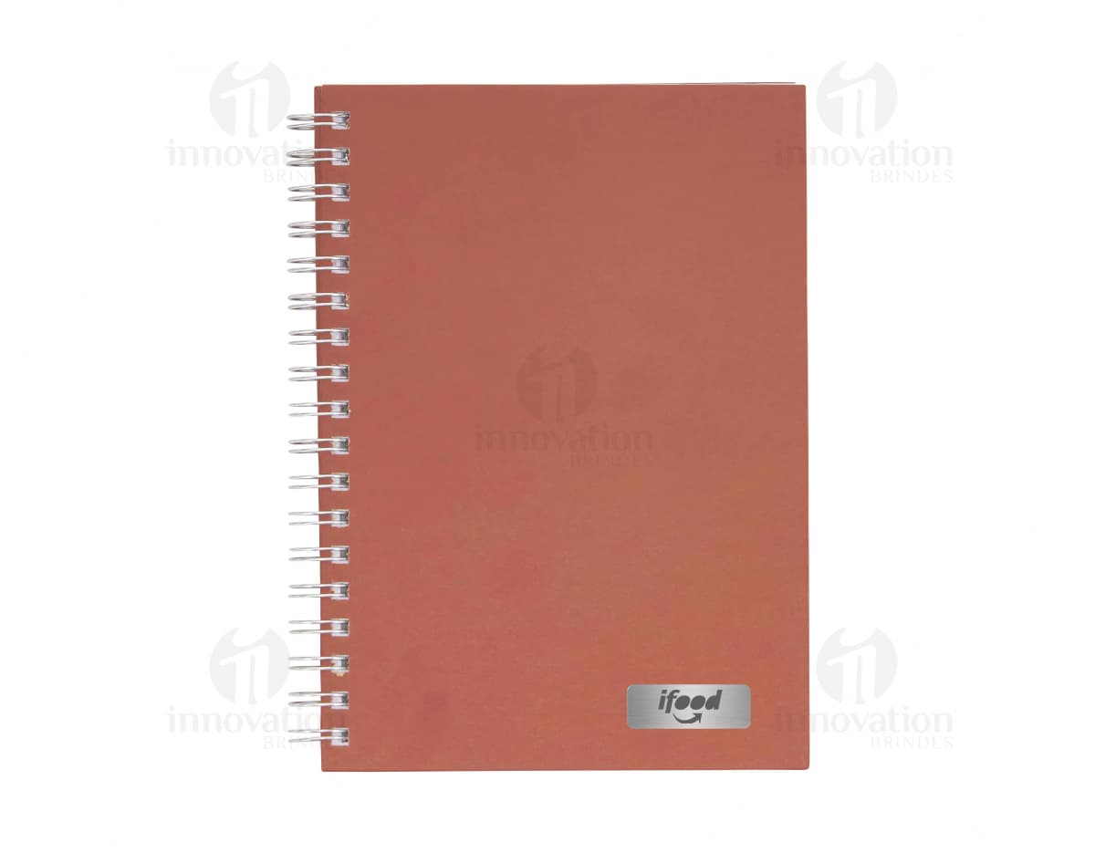 Caderno em branco aberto sobre uma mesa de escritório, com uma caneta ao lado. O caderno possui linhas para escrever e um design simples e moderno. A imagem também mostra um notebook e outros materiais de escrita ao fundo.