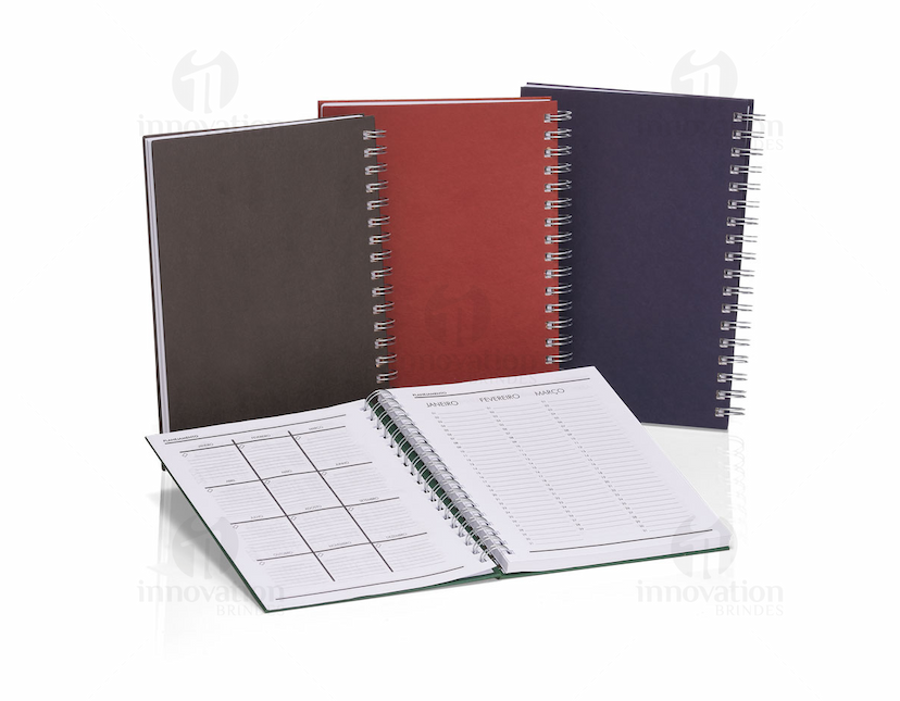 Caderno em branco aberto sobre uma mesa de escritório, com uma caneta ao lado. O caderno possui linhas para escrever e um design simples e moderno. A imagem também mostra um notebook e outros materiais de escrita ao fundo.