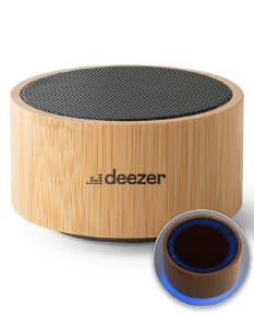 : Caixa de som de bambu e ABS com Bluetooth versão 5.0 em close-up, sobre um bloco de corte. O objeto é metálico, de cor marrom e possui empilhados muffins e cupcakes, com cobertura de chocolate e creme. O dispositivo é brilhante e reflete a luz, indicando alta qualidade.