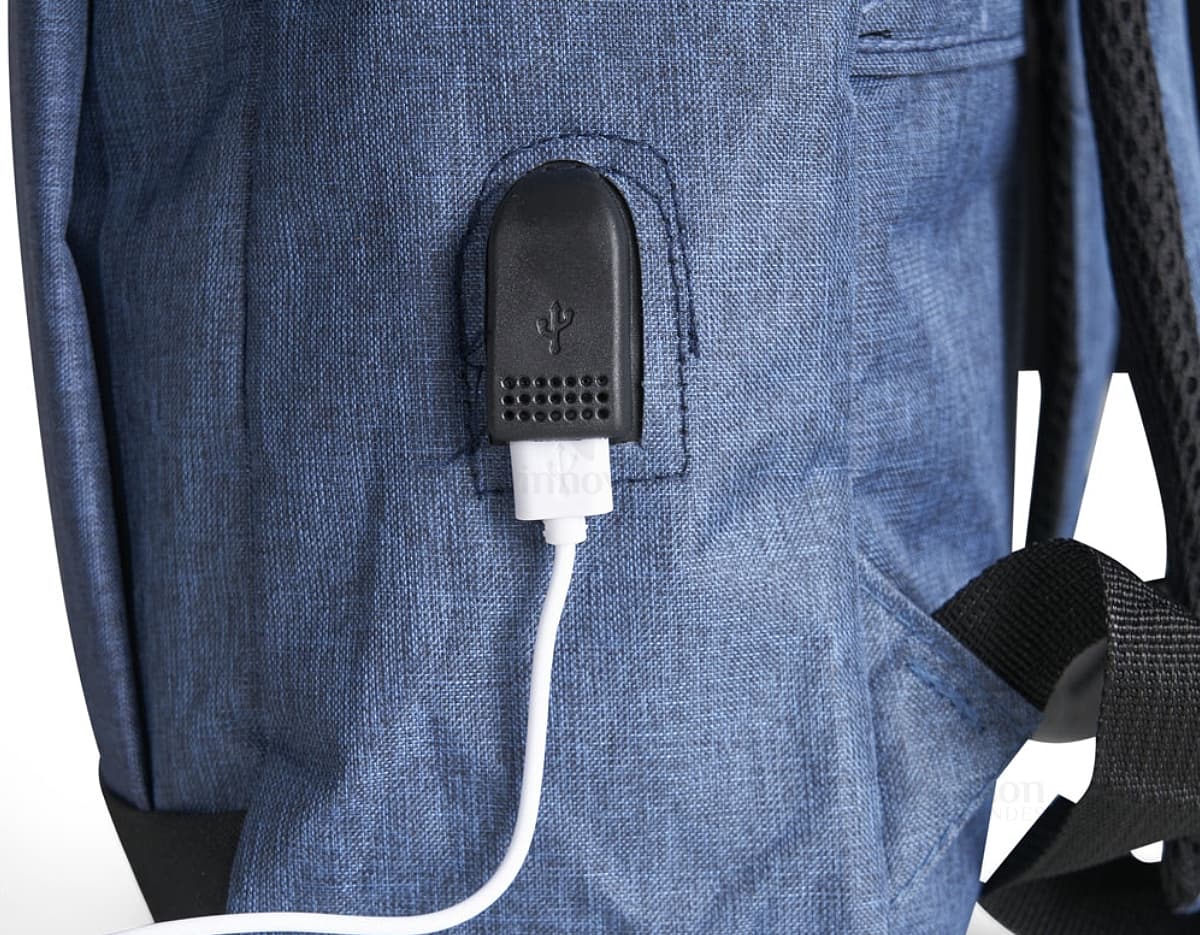 Homem de negócios usando um dispositivo eletrônico em um avião, com uma mochila de nylon preta nas costas. O bolso da mochila contém um cabo USB e há um jeans preto amarrado ao redor da alça.