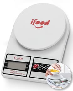 Mulher sorrindo e trabalhando em sua cozinha, com uma mesa de café da manhã com comida, xícara de café, pratos e utensílios. Ela está segurando uma balança digital para cozinha.