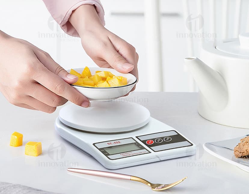 Mulher sorrindo e trabalhando em sua cozinha, com uma mesa de café da manhã com comida, xícara de café, pratos e utensílios. Ela está segurando uma balança digital para cozinha.