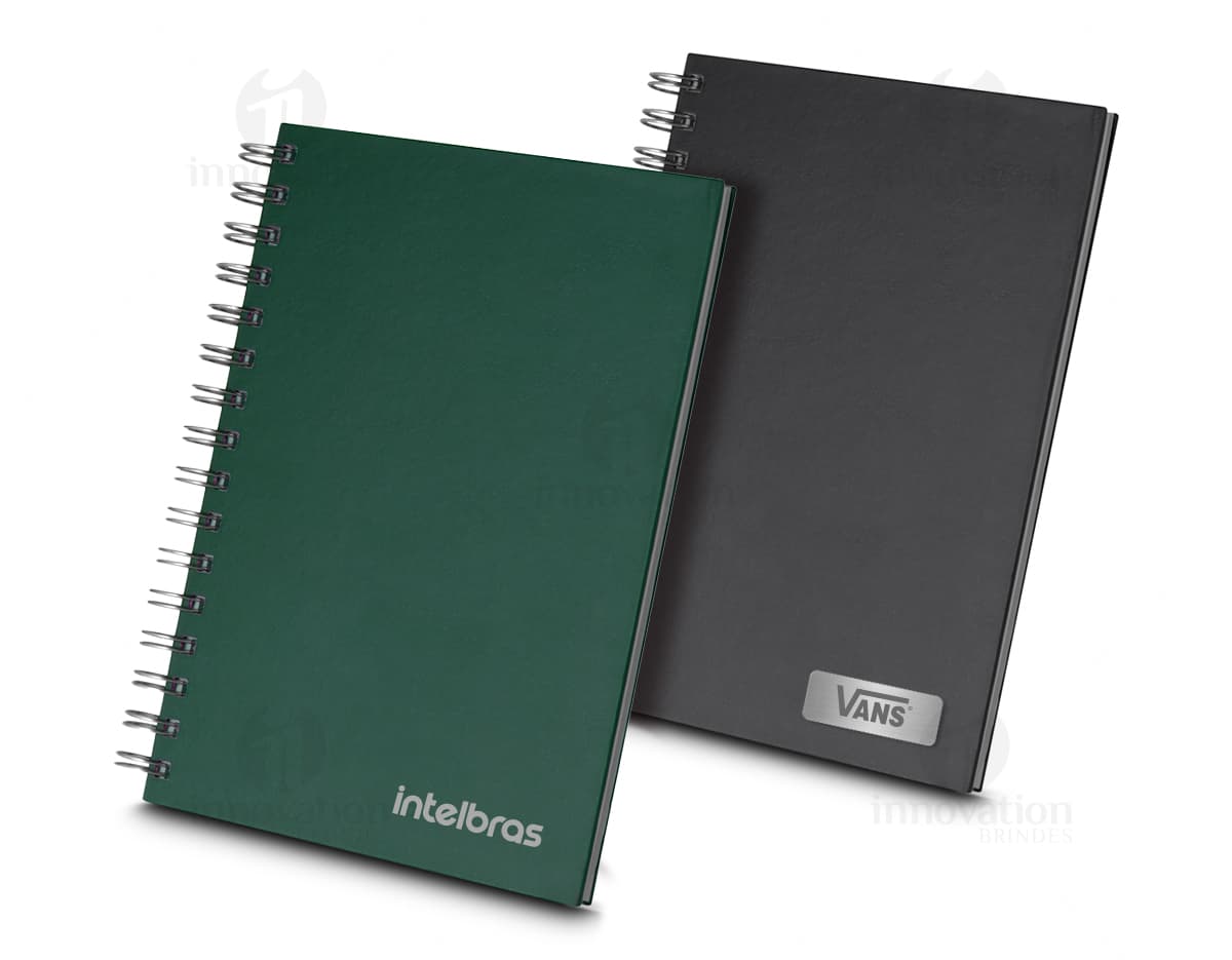Caderno de capa dura preto aberto, com folhas em branco e um lápis sobre ele. Há também um papel e um fichário ao lado. A cena se passa em um ambiente de escritório vazio.