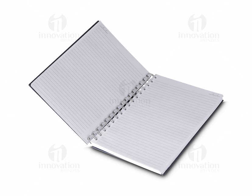 Caderno de capa dura preto aberto, com folhas em branco e um lápis sobre ele. Há também um papel e um fichário ao lado. A cena se passa em um ambiente de escritório vazio.
