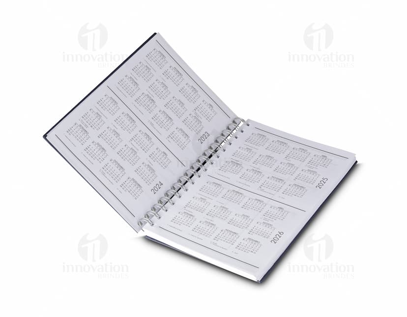 Caderno de capa dura preto aberto, com folhas em branco e um lápis sobre ele. Há também um papel e um fichário ao lado. A cena se passa em um ambiente de escritório vazio.