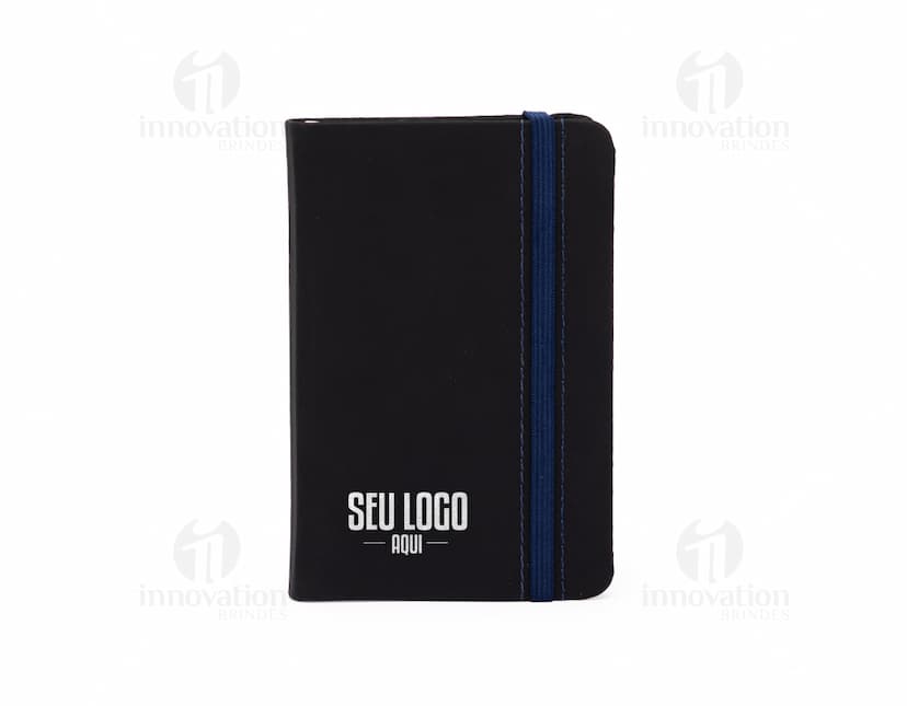 Notebook preto com external drive ao lado, sobre a mesa do escritório. A caderneta emborrachada sem pauta está aberta ao lado do laptop, pronta para anotações. No ambiente de trabalho, diversos objetos de tecnologia e papelaria podem ser vistos em destaque.