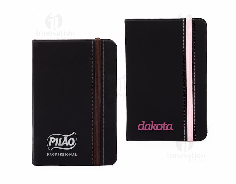 Notebook business preto em branco, com capa de couro e teclado visível. Ao lado há uma carteira de couro preto, além de livros, papéis e uma pasta de documentos. No teclado do notebook, há uma carteira aberta com dinheiro e uma nota. A imagem inclui também um estojo de escritório, um ícone de cifrão, botões de teclado e um alto-falante.