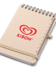 Mini caderno em 3D, com capa preta e espiral, aberto e vazio. O caderno está sobre uma mesa de escritório, ao lado de um notebook, um livro e um papel em branco. A imagem transmite a ideia de organização, estudo e trabalho.