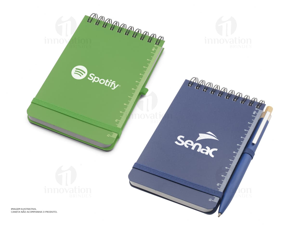 Mini caderno em 3D, com capa preta e espiral, aberto e vazio. O caderno está sobre uma mesa de escritório, ao lado de um notebook, um livro e um papel em branco. A imagem transmite a ideia de organização, estudo e trabalho.