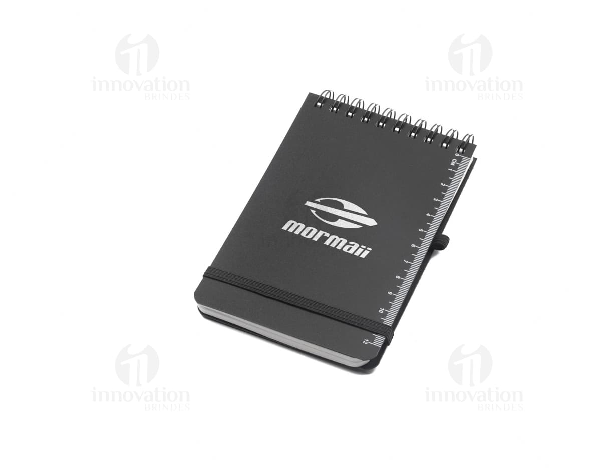 Mini caderno em 3D, com capa preta e espiral, aberto e vazio. O caderno está sobre uma mesa de escritório, ao lado de um notebook, um livro e um papel em branco. A imagem transmite a ideia de organização, estudo e trabalho.
