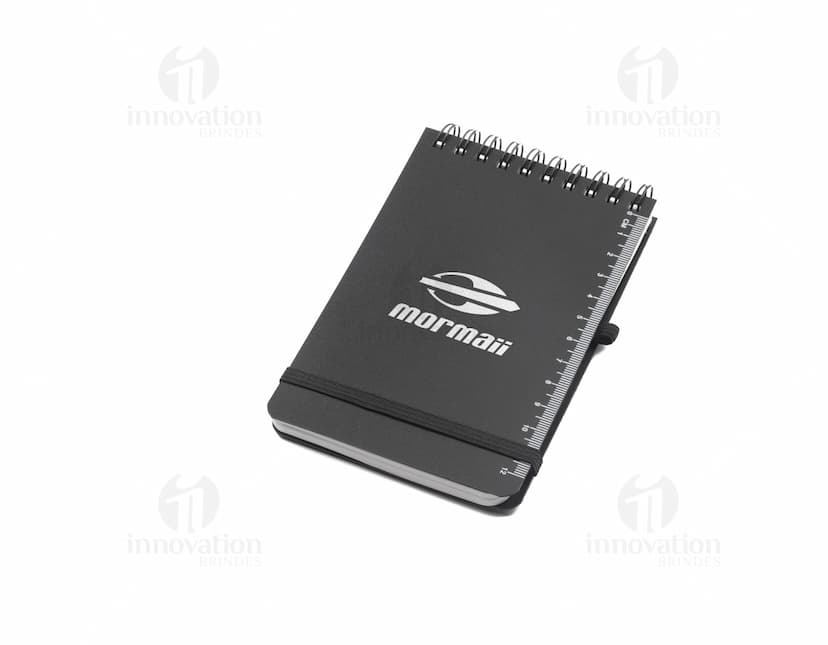 Mini caderno em 3D, com capa preta e espiral, aberto e vazio. O caderno está sobre uma mesa de escritório, ao lado de um notebook, um livro e um papel em branco. A imagem transmite a ideia de organização, estudo e trabalho.