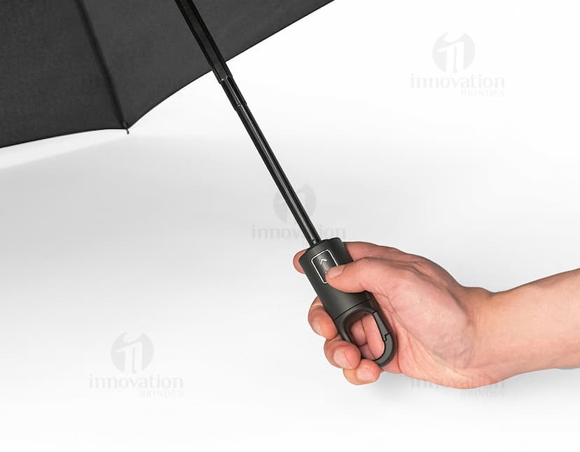 Guarda-chuva dobrável preto em formato de capelo de formatura, simbolizando sucesso e educação. O objeto 3D está aberto e vazio, sendo segurado por uma mão masculina. A imagem representa conhecimento, celebração e conquista acadêmica.