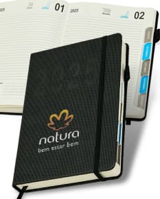Agenda Diária 2025 em couro marrom, aberta sobre um notebook preto com papel em branco, teclado de computador e diversos objetos de escritório ao redor, como caneta, carteira, pasta e dinheiro.