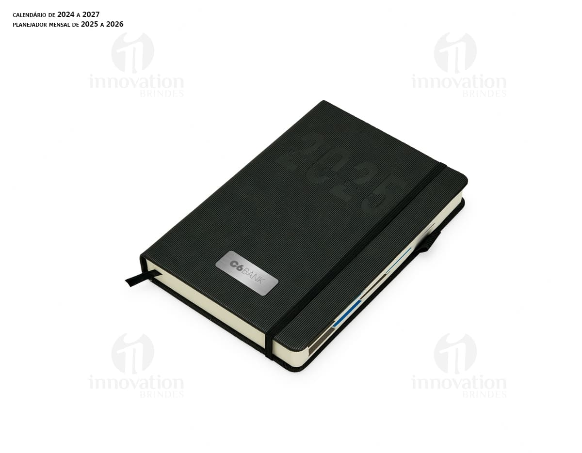 Agenda Diária 2025 em couro marrom, aberta sobre um notebook preto com papel em branco, teclado de computador e diversos objetos de escritório ao redor, como caneta, carteira, pasta e dinheiro.