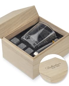 Kit Drink em uma caixa de papelão marrom. A embalagem está aberta e contém várias garrafas e copos empilhados. Ao redor da caixa, há diversos objetos de decoração e símbolos de riqueza, como um cofre, livros, um baú e uma impressora.