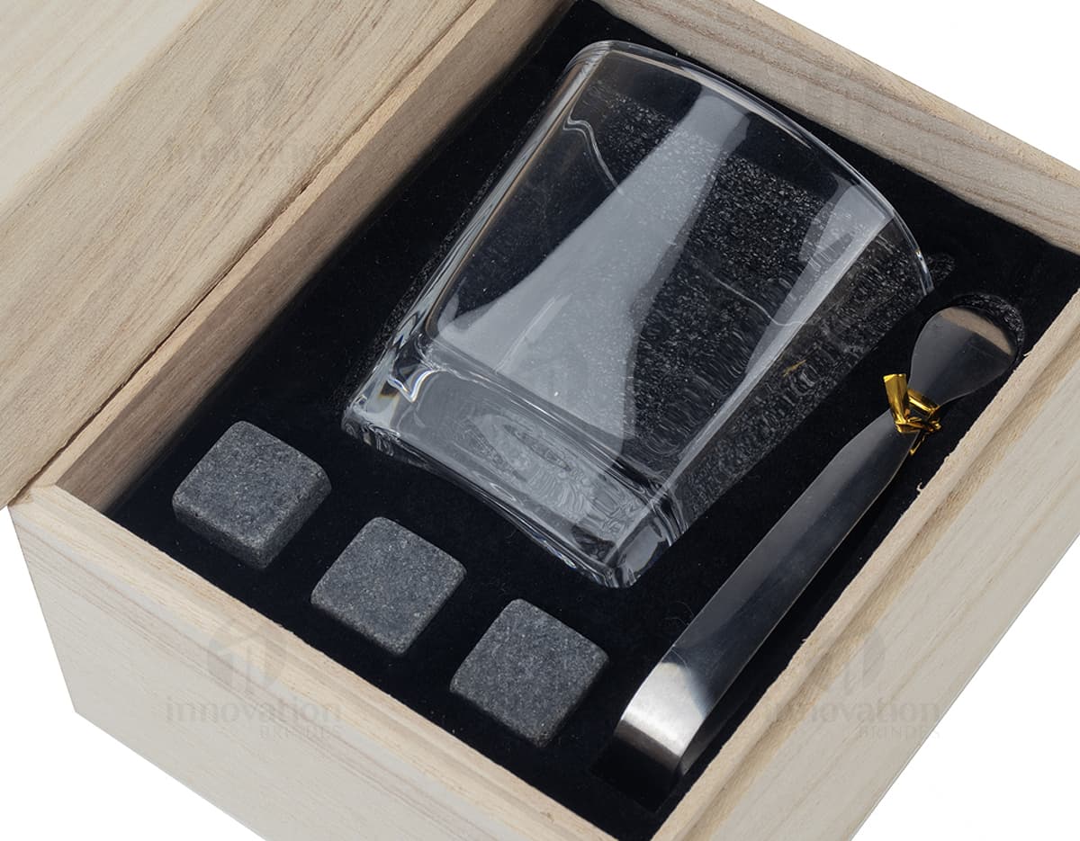 Kit Drink em uma caixa de papelão marrom. A embalagem está aberta e contém várias garrafas e copos empilhados. Ao redor da caixa, há diversos objetos de decoração e símbolos de riqueza, como um cofre, livros, um baú e uma impressora.