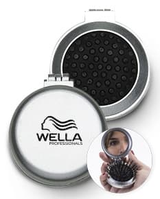 Espelho de bolsa com escova preta e vermelha, feito de vidro e metal, com um botão redondo para fechar. Possui um ícone de lupa prateada, símbolo de pesquisa e ampliação. O objeto é um instrumento de maquiagem compacto e prático para levar na bolsa, ideal para retoques durante o dia.