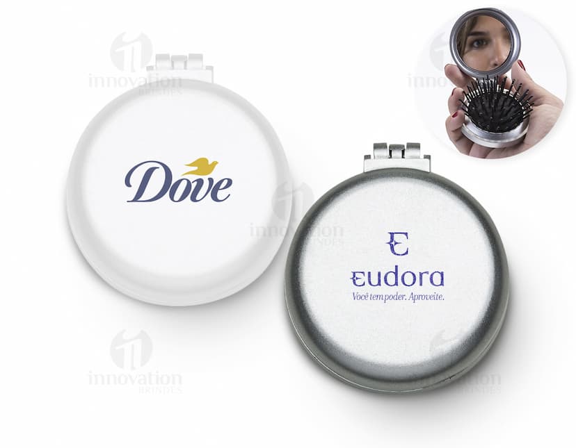 Espelho de bolsa com escova preta e vermelha, feito de vidro e metal, com um botão redondo para fechar. Possui um ícone de lupa prateada, símbolo de pesquisa e ampliação. O objeto é um instrumento de maquiagem compacto e prático para levar na bolsa, ideal para retoques durante o dia.