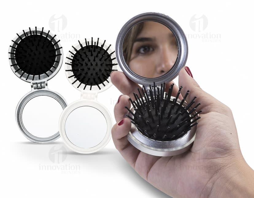 Espelho de bolsa com escova preta e vermelha, feito de vidro e metal, com um botão redondo para fechar. Possui um ícone de lupa prateada, símbolo de pesquisa e ampliação. O objeto é um instrumento de maquiagem compacto e prático para levar na bolsa, ideal para retoques durante o dia.