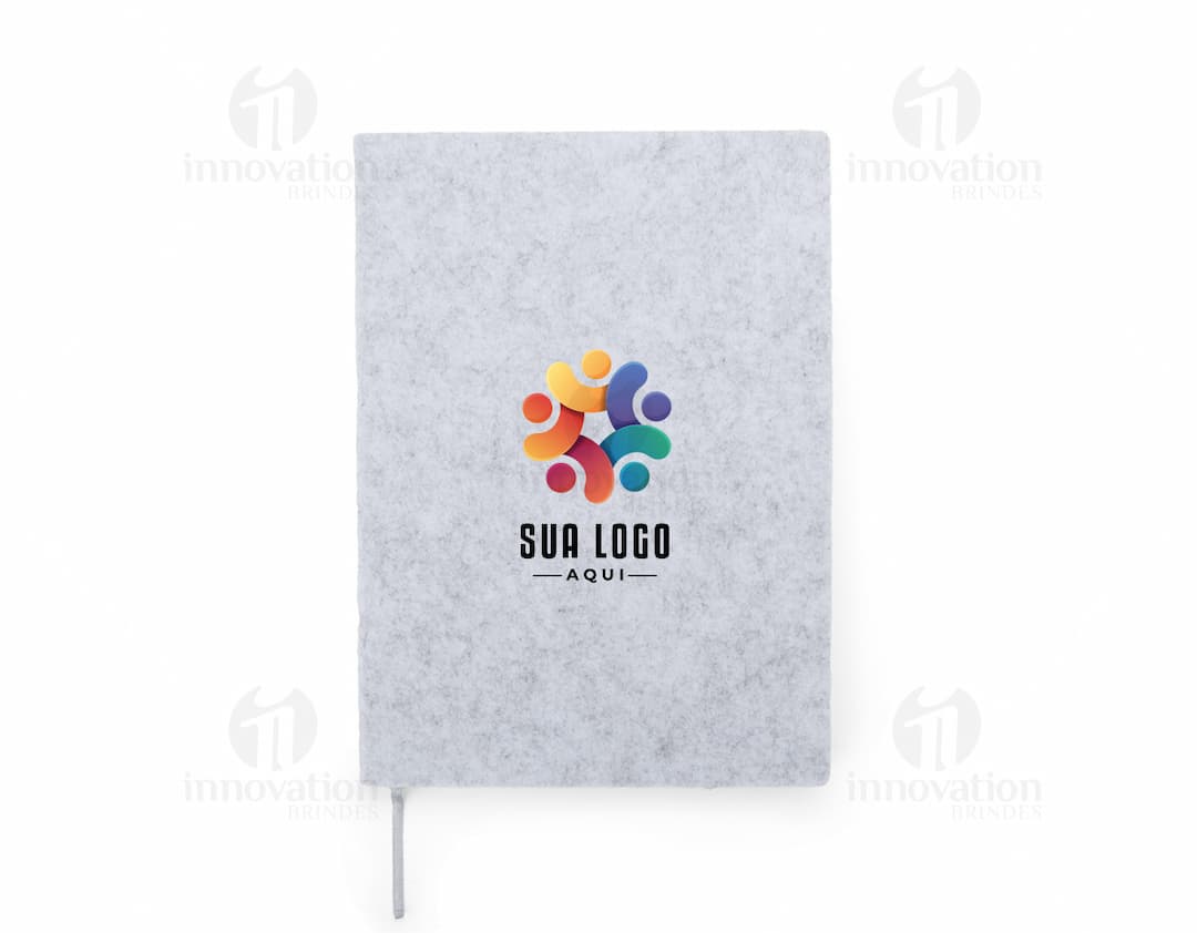 caderno A5 de feltro preto, sem pauta, aberto em uma página em branco. Ao lado, um notebook e um envelope preto. No fundo, livros e objetos de escritório.