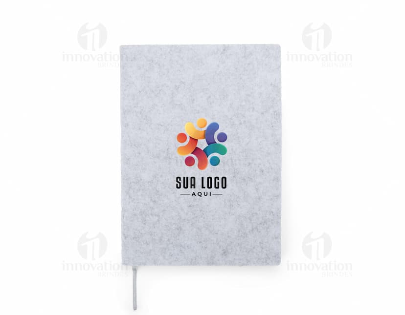 caderno A5 de feltro preto, sem pauta, aberto em uma página em branco. Ao lado, um notebook e um envelope preto. No fundo, livros e objetos de escritório.