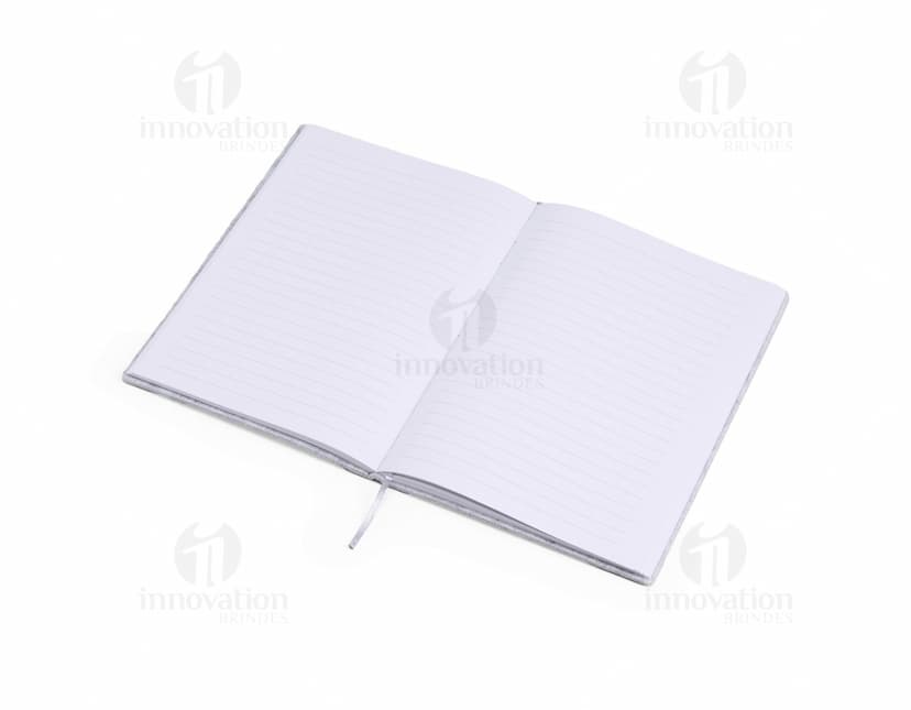 caderno A5 de feltro preto, sem pauta, aberto em uma página em branco. Ao lado, um notebook e um envelope preto. No fundo, livros e objetos de escritório.