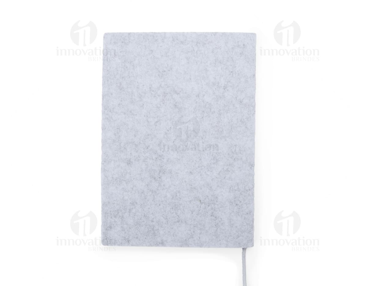caderno A5 de feltro preto, sem pauta, aberto em uma página em branco. Ao lado, um notebook e um envelope preto. No fundo, livros e objetos de escritório.