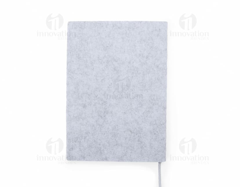 caderno A5 de feltro preto, sem pauta, aberto em uma página em branco. Ao lado, um notebook e um envelope preto. No fundo, livros e objetos de escritório.