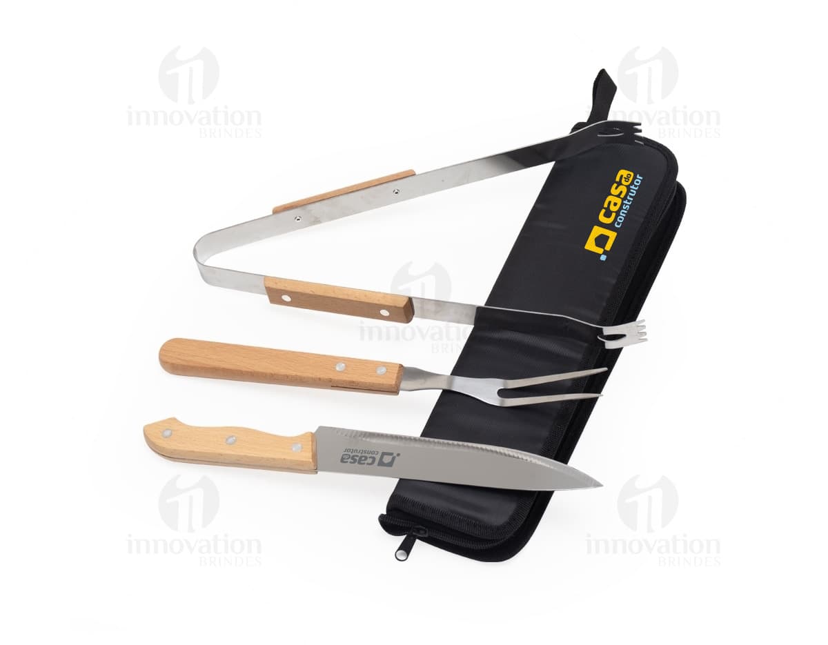 Kit churrasco composto por 3 peças: faca, garfo e pegador de carne em aço inoxidável, sobre fundo de madeira.