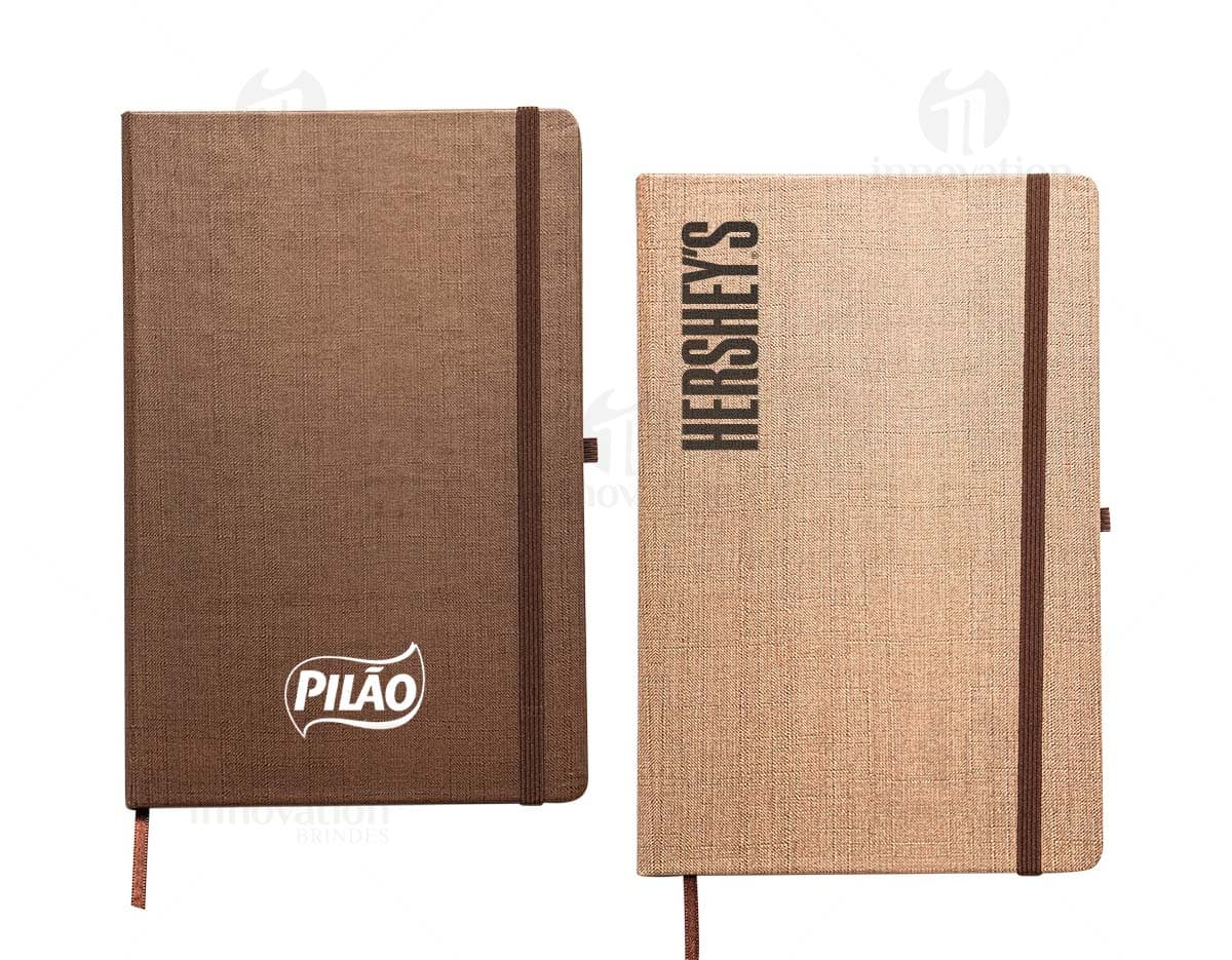 Caderneta A5 em papel texturizado antigo, com capa de tecido vintage em tom de marrom desgastado. O design é grunge e possui um padrão decorativo. O material é de fibra de algodão áspera, com uma superfície em canvas. Na capa, há um detalhe de costura em linha preta. O interior está em branco, com páginas vazias.