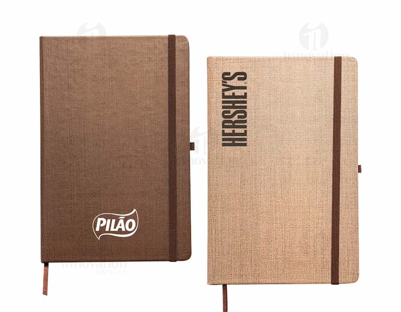 Caderneta A5 em papel texturizado antigo, com capa de tecido vintage em tom de marrom desgastado. O design é grunge e possui um padrão decorativo. O material é de fibra de algodão áspera, com uma superfície em canvas. Na capa, há um detalhe de costura em linha preta. O interior está em branco, com páginas vazias.