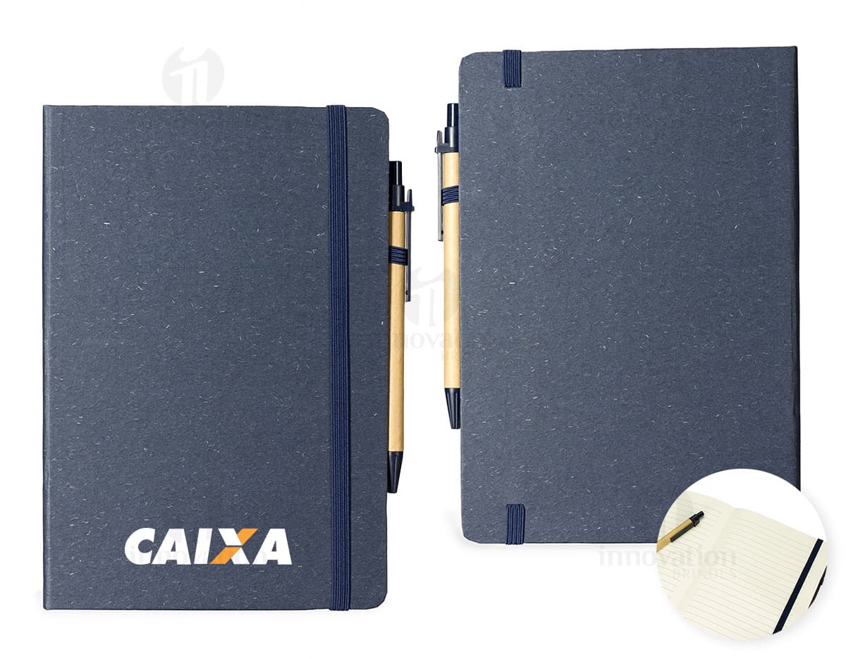 Caderneta de fibra de arroz com caneta em fundo branco. O caderno é marrom claro e possui detalhes em preto na capa. A caneta é de cor azul e está ao lado da caderneta.