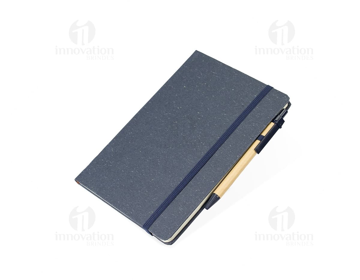 Caderneta de fibra de arroz com caneta em fundo branco. O caderno é marrom claro e possui detalhes em preto na capa. A caneta é de cor azul e está ao lado da caderneta.