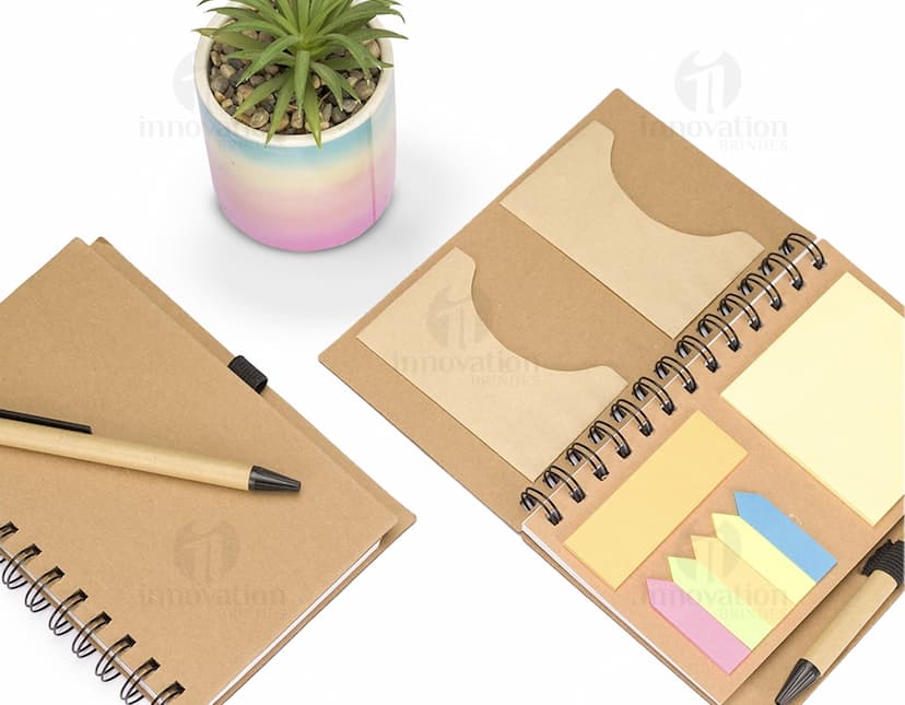 Caderno Ecológico com capa de papel kraft marrom, fecho de elástico e detalhe de folha verde. Páginas internas em papel reciclado.
