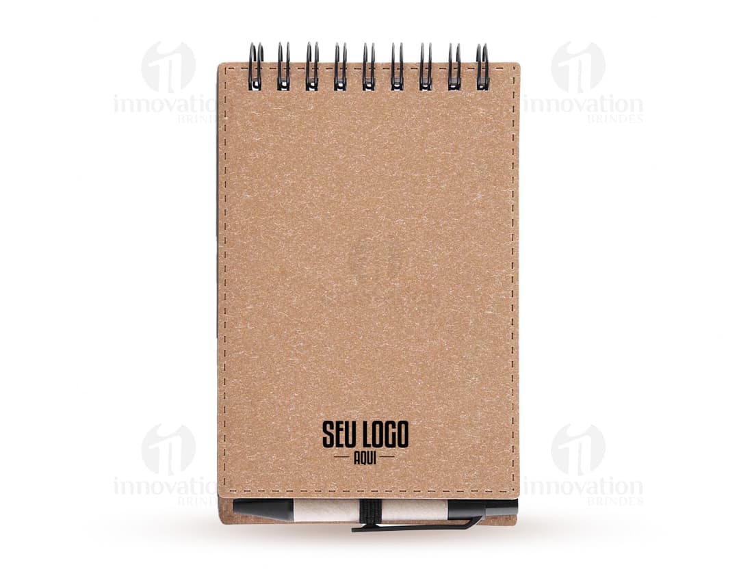 Mini caderno de anotações em papel em branco, com capa marrom de cartão. Acompanha uma caneta e está em cima de um notebook