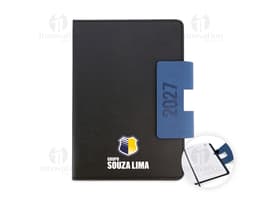 Notebook preto aberto, com teclado e tela exibindo ícones e botões. Ao lado, uma agenda A5 com capa dura em PU, destacando-se