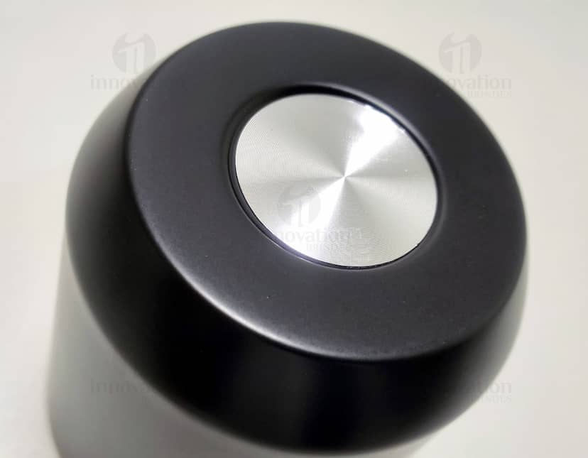 Copo térmico de 280ml em cerâmica preta e metal de cor prateada, com alça e tampa. O copo está vazio e posicionado sobre uma mesa de cozinha, ao lado de uma colher, uma jarra de café e alguns utensílios.