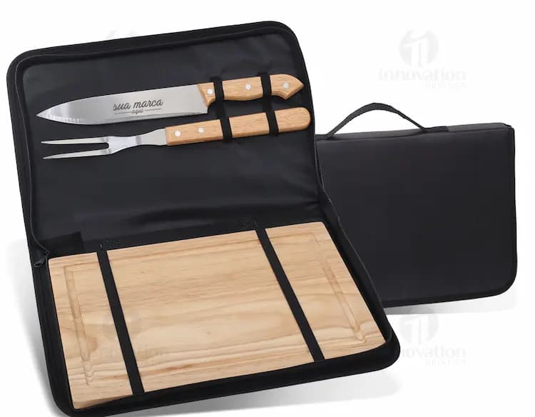 Kit queijo 3 peças em um estojo de kraft com orifício, contendo uma faca, uma espátula e um garfo para queijo. O kit está posicionado em cima de uma tábua de madeira, ao lado de um monte de nozes, amêndoas e frutas secas. A imagem transmite uma sensação de gourmet e saúde, com tons de marrom e amarelo predominantes.